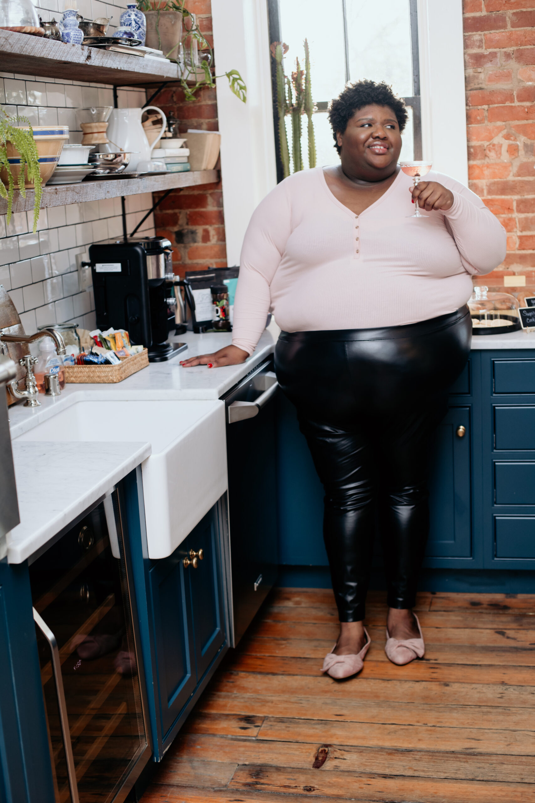 Kandice is standing in a kitchen nook, holding a pink cocktail.