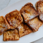 Brioche french toast sitting on a platter, drizzled with male syrup and dusted with powdered sugar.