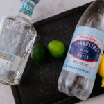 Tequila, limes, and sparkling water are displayed on a cutting board.